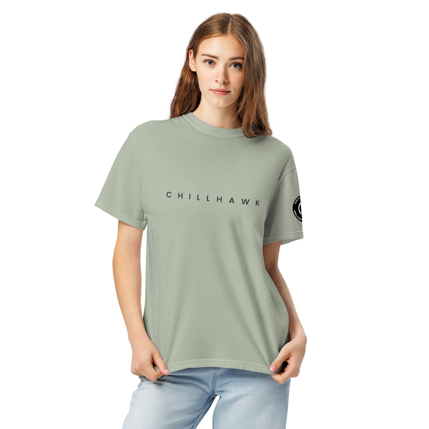 Chillhawk unisex heavyweight short sleeve t-shirt in bay - a lady with light skin and long brown hair, is
wearing the t-shirt and light blue jeans. The word "CHILLHAWK" is printed in black on the front of the shirt, and a circular logo is visible on the left sleeve.
