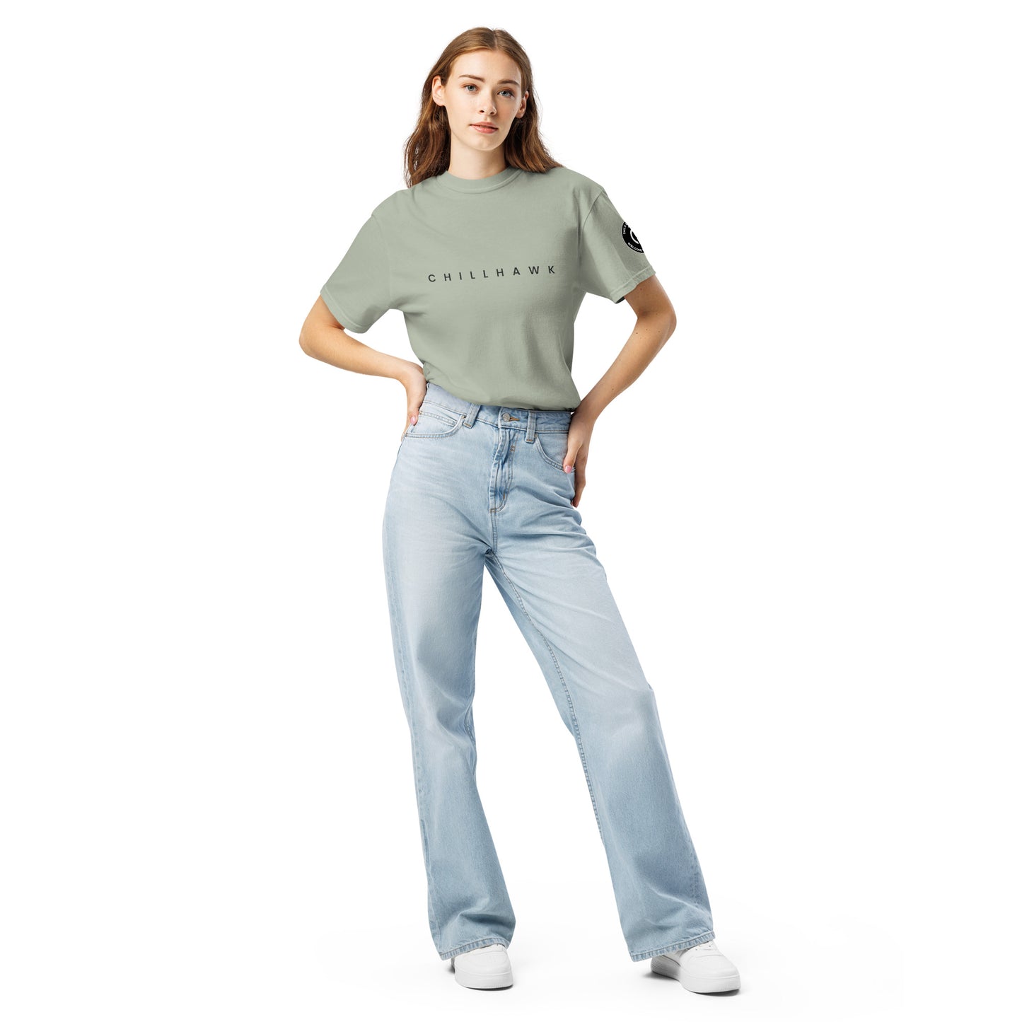 Chillhawk unisex heavyweight short sleeve t-shirt in bay - a lady with light skin and long brown hair, is standing with hands on hips wearing the t-shirt, light blue jeans, and white sneakers. The word "CHILLHAWK" is printed in black on the front of the shirt, and a circular logo is visible on the left sleeve.