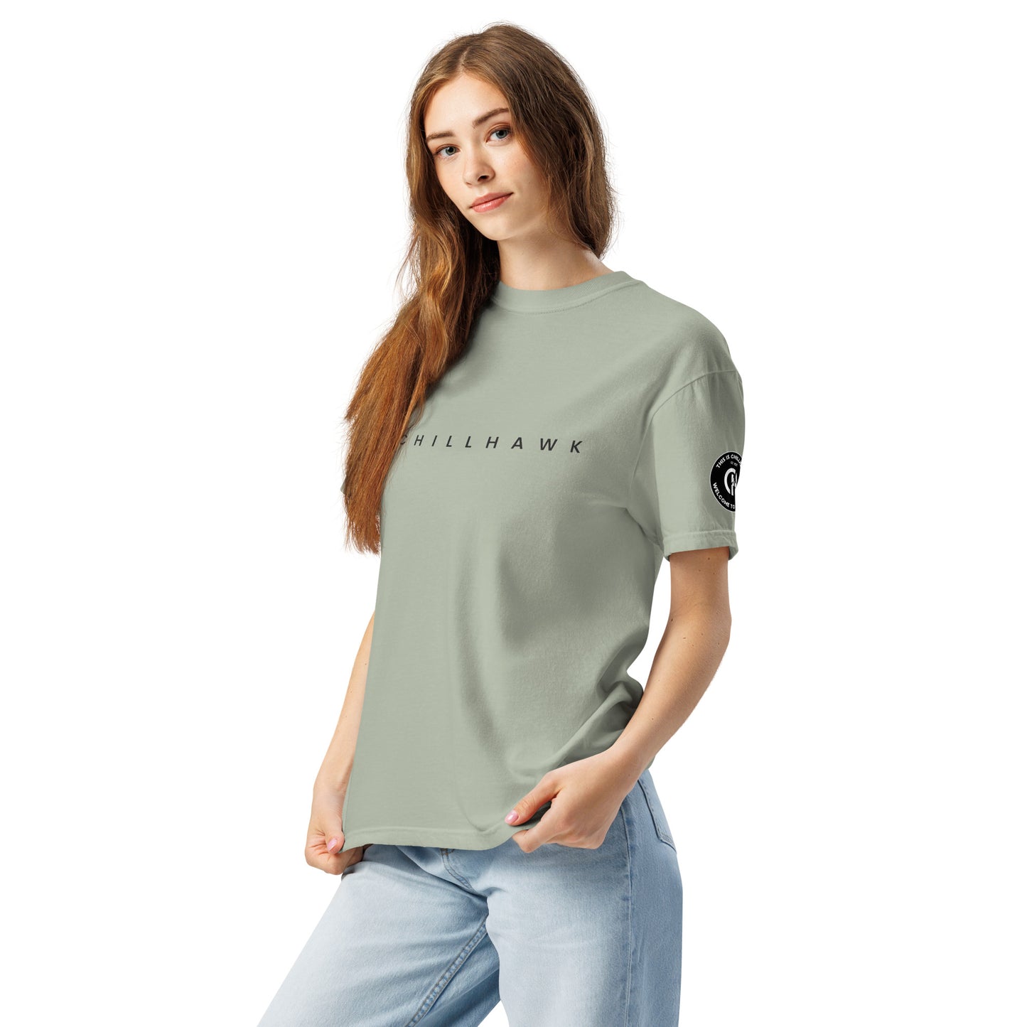 Chillhawk unisex heavyweight short sleeve t-shirt in bay - a lady with light skin and long brown hair, is
wearing the t-shirt and light blue jeans. The word "CHILLHAWK" is printed in black on the front of the shirt, and a circular logo is visible on the left sleeve.