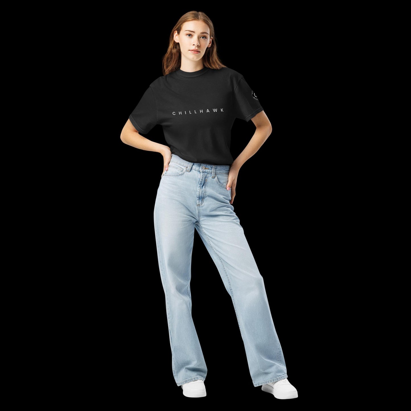 Chillhawk unisex heavyweight short sleeve t-shirt in black - front view of a lady with light skin and long brown hair, is wearing the t-shirt and light blue jeans. The word "CHILLHAWK" is printed in black on the front of the shirt, and a circular logo is visible on the left sleeve.