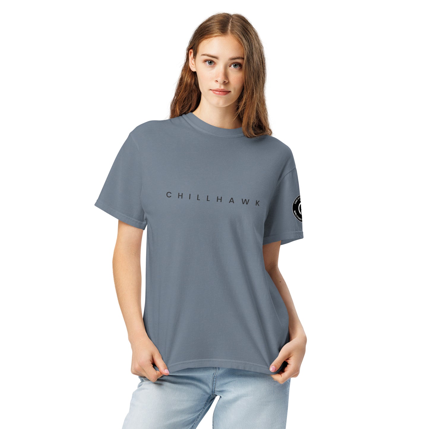 Chillhawk unisex heavyweight short sleeve t-shirt in slate blue - a lady with light skin and long brown hair, is
wearing the t-shirt and light blue jeans. The word "CHILLHAWK" is printed in black on the front of the shirt, and a circular logo is visible on the left sleeve.