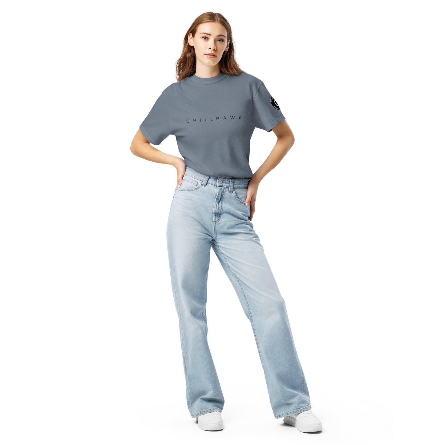 Chillhawk unisex heavyweight short sleeve t-shirt in slate blue - a lady with light skin and long brown hair, is standing with hands on hips wearing the t-shirt, light blue jeans, and white sneakers. The word "CHILLHAWK" is printed in black on the front of the shirt, and a circular logo is visible on the left sleeve.