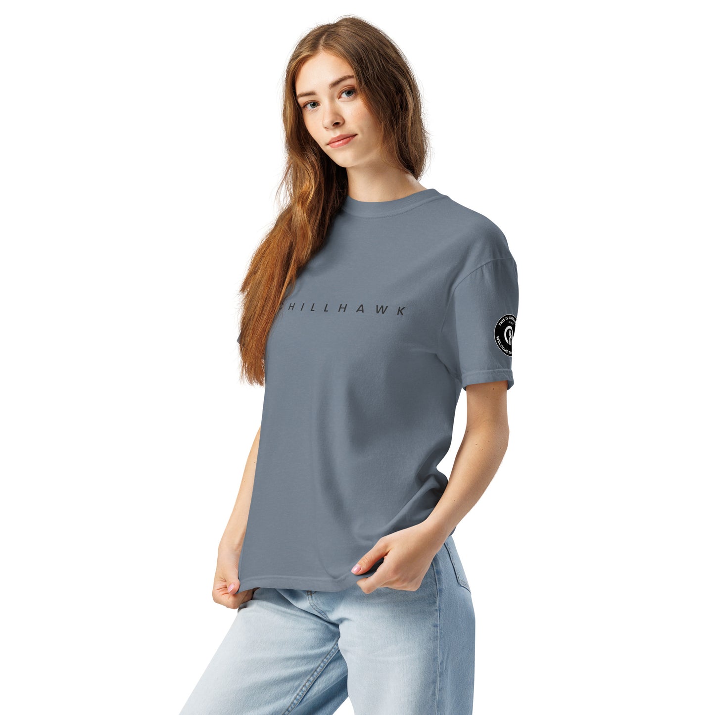 Chillhawk unisex heavyweight short sleeve t-shirt in slate blue - a lady with light skin and long brown hair, is wearing the t-shirt and light blue jeans. The word "CHILLHAWK" is printed in black on the front of the shirt, and a circular logo is visible on the left sleeve.