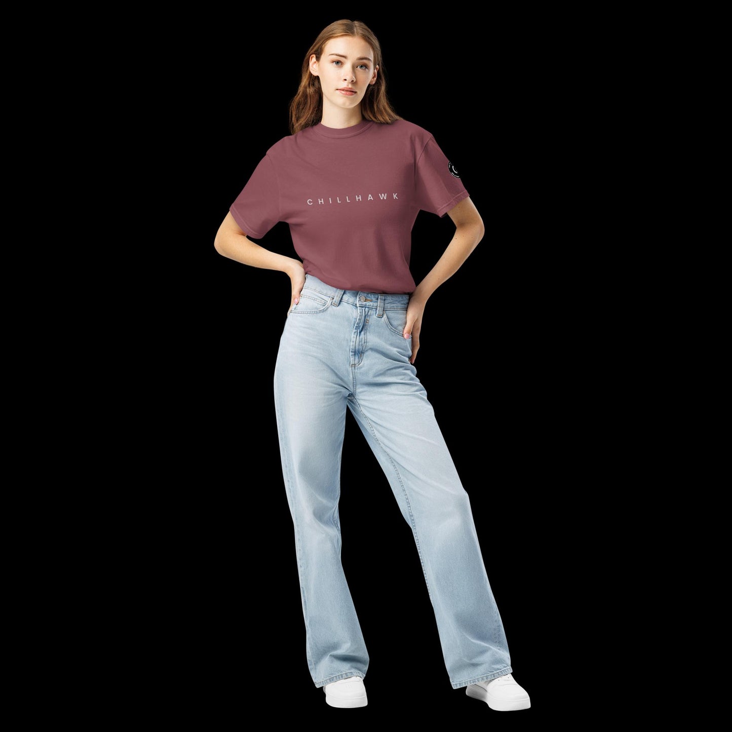 Chillhawk unisex heavyweight short sleeve t-shirt in brick - front view of a lady with light skin and long brown hair, is wearing the t-shirt and light blue jeans. The word "CHILLHAWK" is printed in black on the front of the shirt, and a circular logo is visible on the left sleeve.