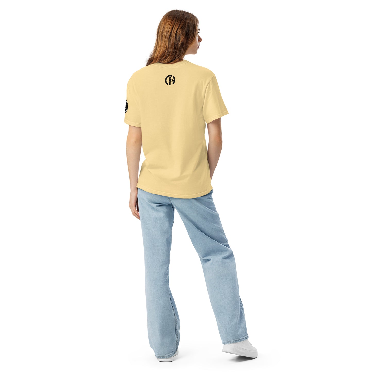 Chillhawk unisex heavyweight short sleeve t-shirt in butter - back view of a lady with light skin and long brown hair, is wearing the t-shirt, light blue jeans, and white sneakers. A circular logo is visible on the back side.