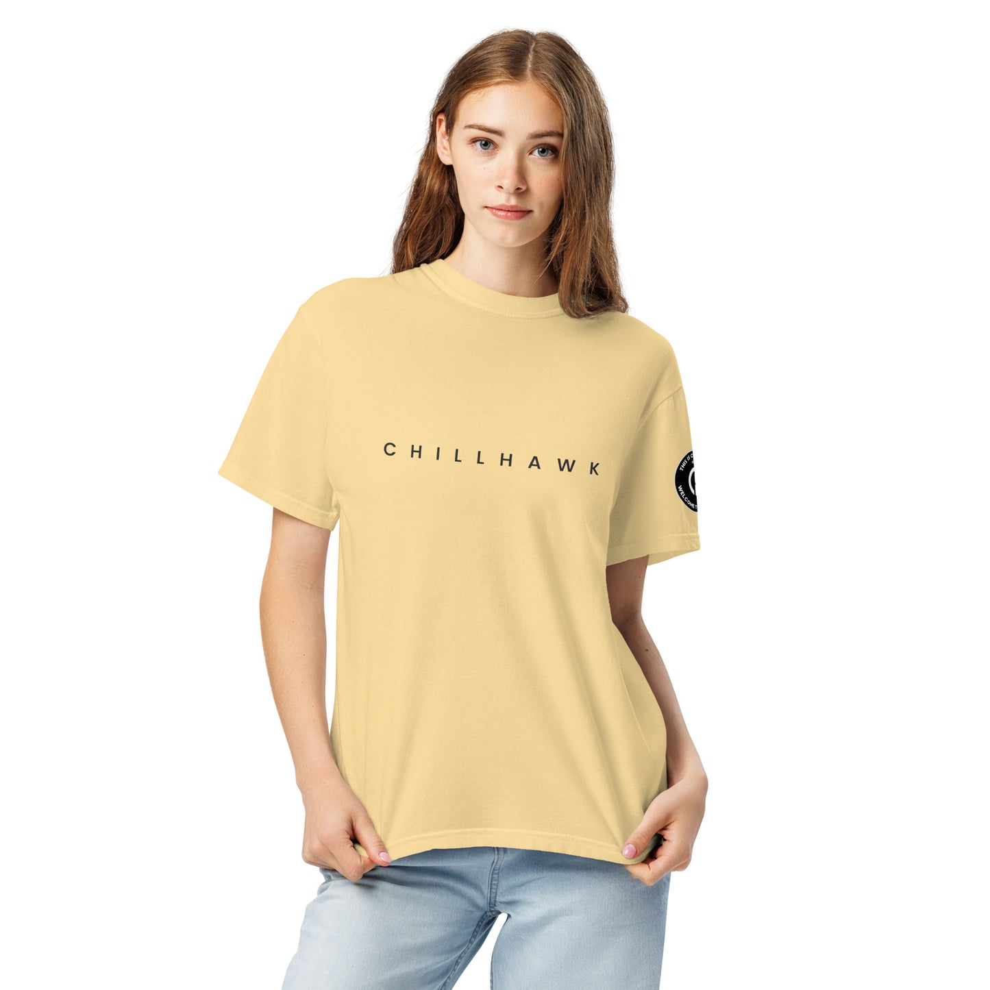 Chillhawk unisex heavyweight short sleeve t-shirt in butter - a lady with light skin and long brown hair, is
wearing the t-shirt and light blue jeans. The word "CHILLHAWK" is printed in black on the front of the shirt, and a circular logo is visible on the left sleeve.