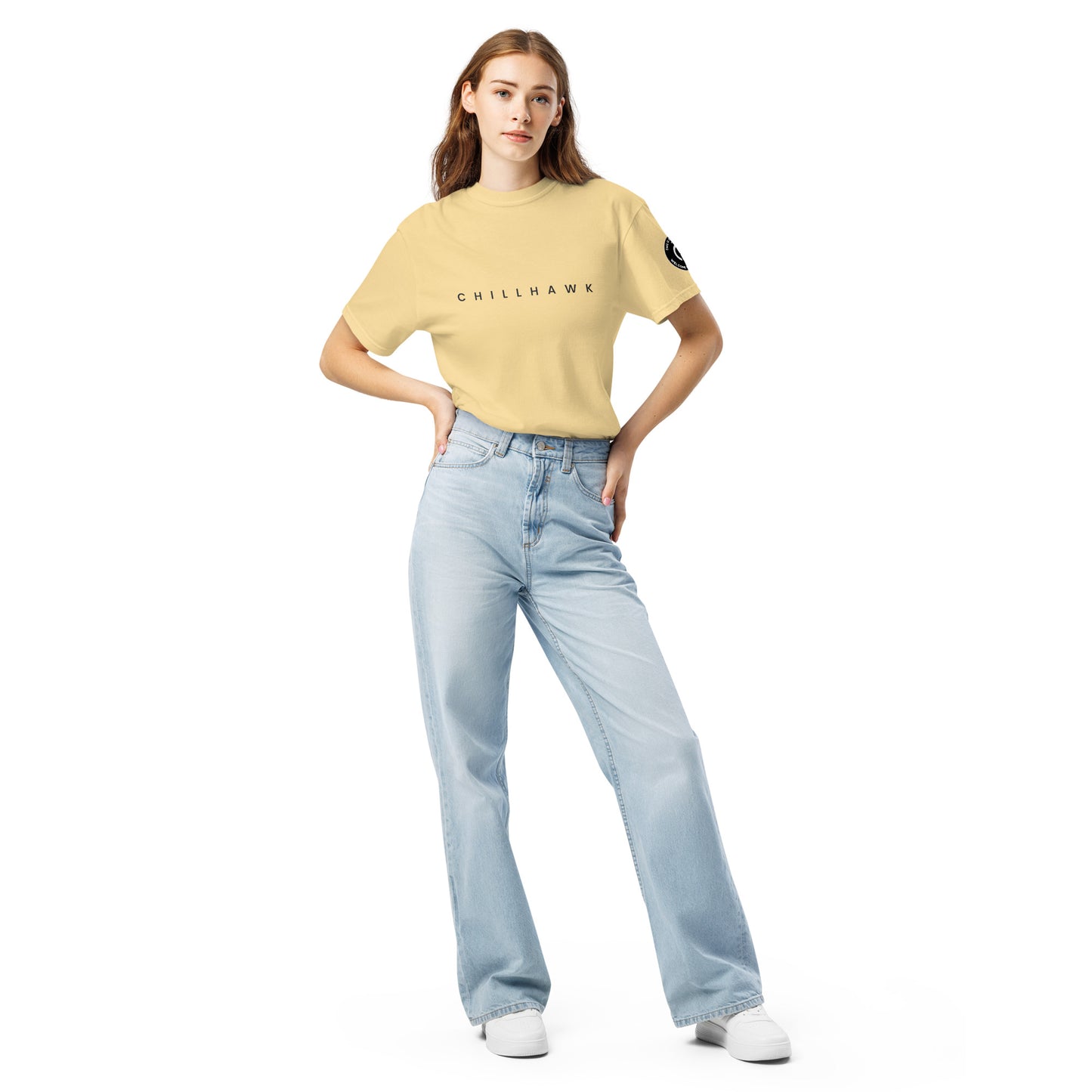 Chillhawk unisex heavyweight short sleeve t-shirt in butter - a lady with light skin and long brown hair, is standing with hands on hips wearing the t-shirt, light blue jeans, and white sneakers. The word "CHILLHAWK" is printed in black on the front of the shirt, and a circular logo is visible on the left sleeve.