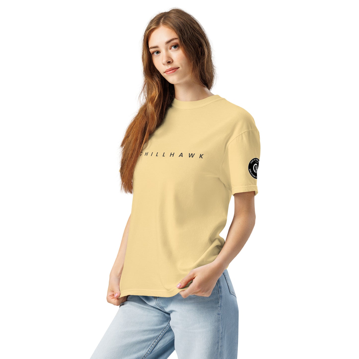 Chillhawk unisex heavyweight short sleeve t-shirt in butter - a lady with light skin and long brown hair, is
wearing the t-shirt and light blue jeans. The word "CHILLHAWK" is printed in black on the front of the shirt, and a circular logo is visible on the left sleeve.
