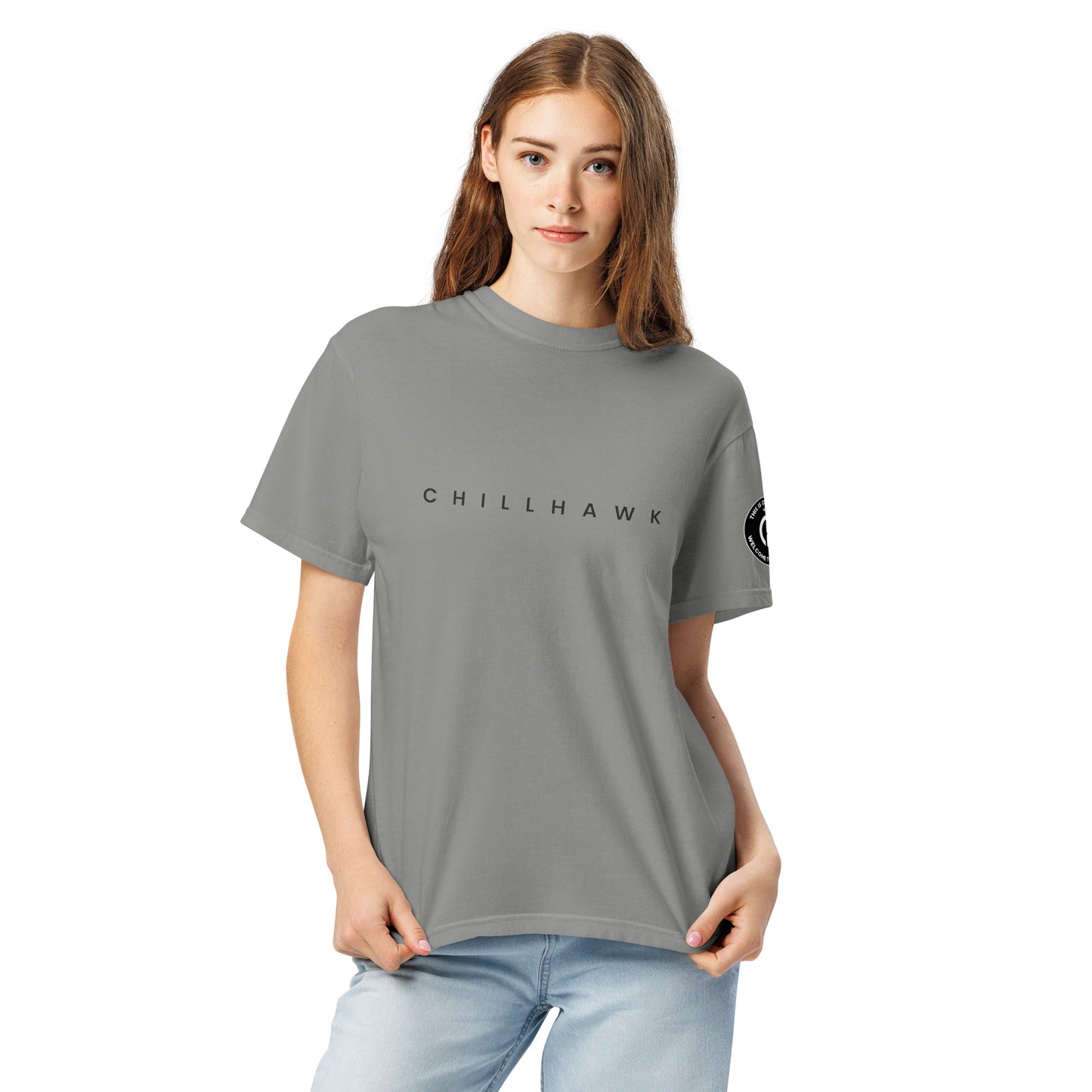 Chillhawk unisex heavyweight short sleeve t-shirt in grey - a lady with light skin and long brown hair, is
wearing the t-shirt and light blue jeans. The word "CHILLHAWK" is printed in black on the front of the shirt, and a circular logo is visible on the left sleeve.