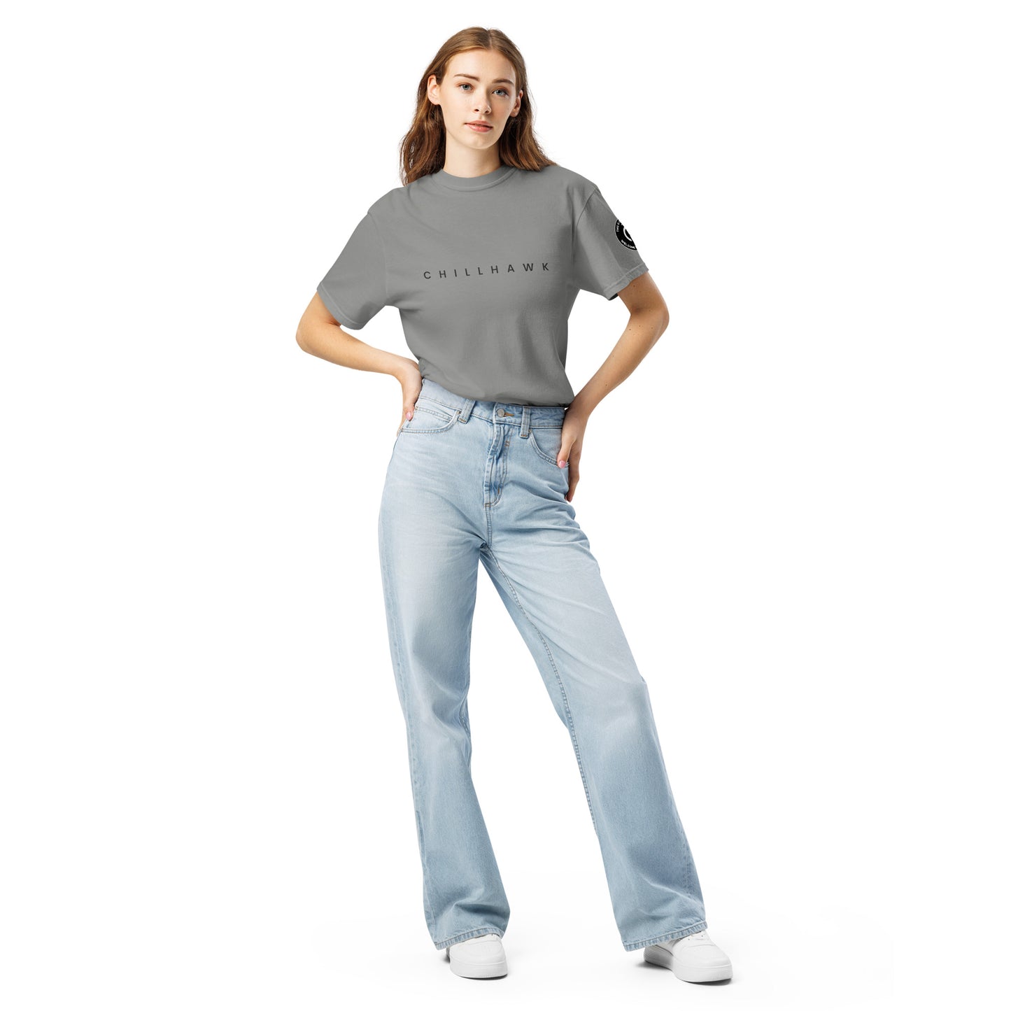 Chillhawk unisex heavyweight short sleeve t-shirt in grey - a lady with light skin and long brown hair, is standing with hands on hips wearing the t-shirt, light blue jeans, and white sneakers. The word "CHILLHAWK" is printed in black on the front of the shirt, and a circular logo is visible on the left sleeve.