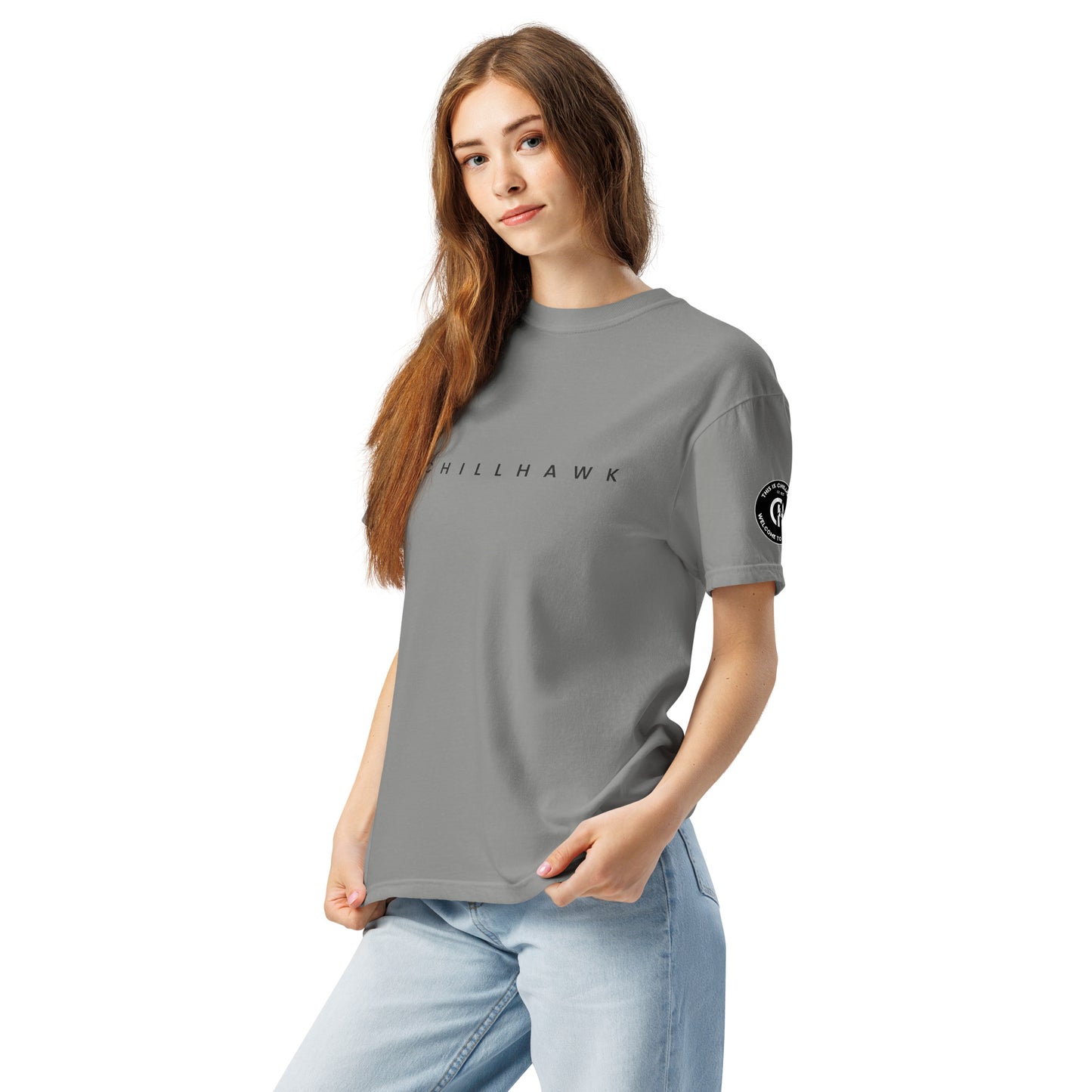 Chillhawk unisex heavyweight short sleeve t-shirt in grey - a lady with light skin and long brown hair, is
wearing the t-shirt and light blue jeans. The word "CHILLHAWK" is printed in black on the front of the shirt, and a circular logo is visible on the left sleeve.