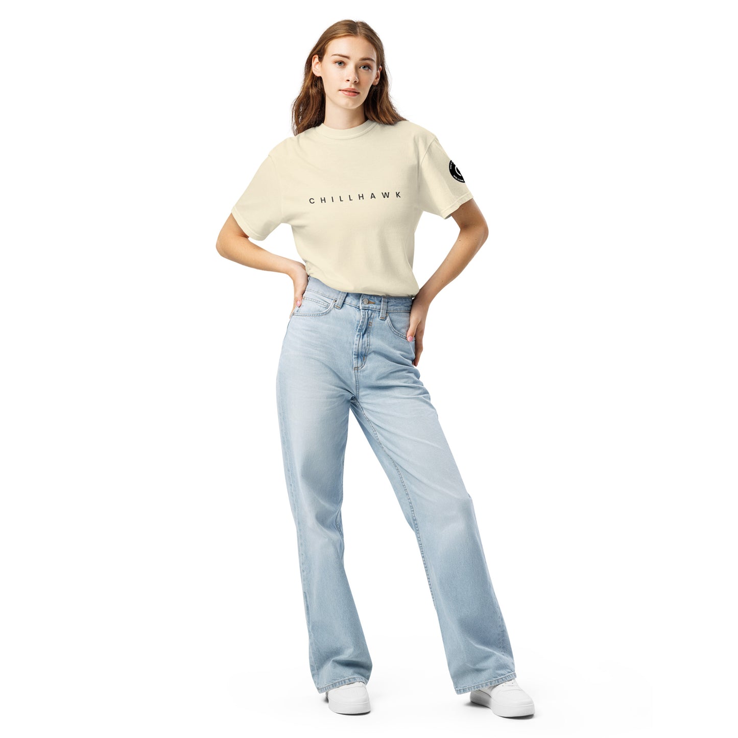Chillhawk unisex heavyweight short sleeve t-shirt in ivory - a lady with light skin and long brown hair, is standing with hands on hips wearing the t-shirt, light blue jeans, and white sneakers. The word "CHILLHAWK" is printed in black on the front of the shirt, and a circular logo is visible on the left sleeve.