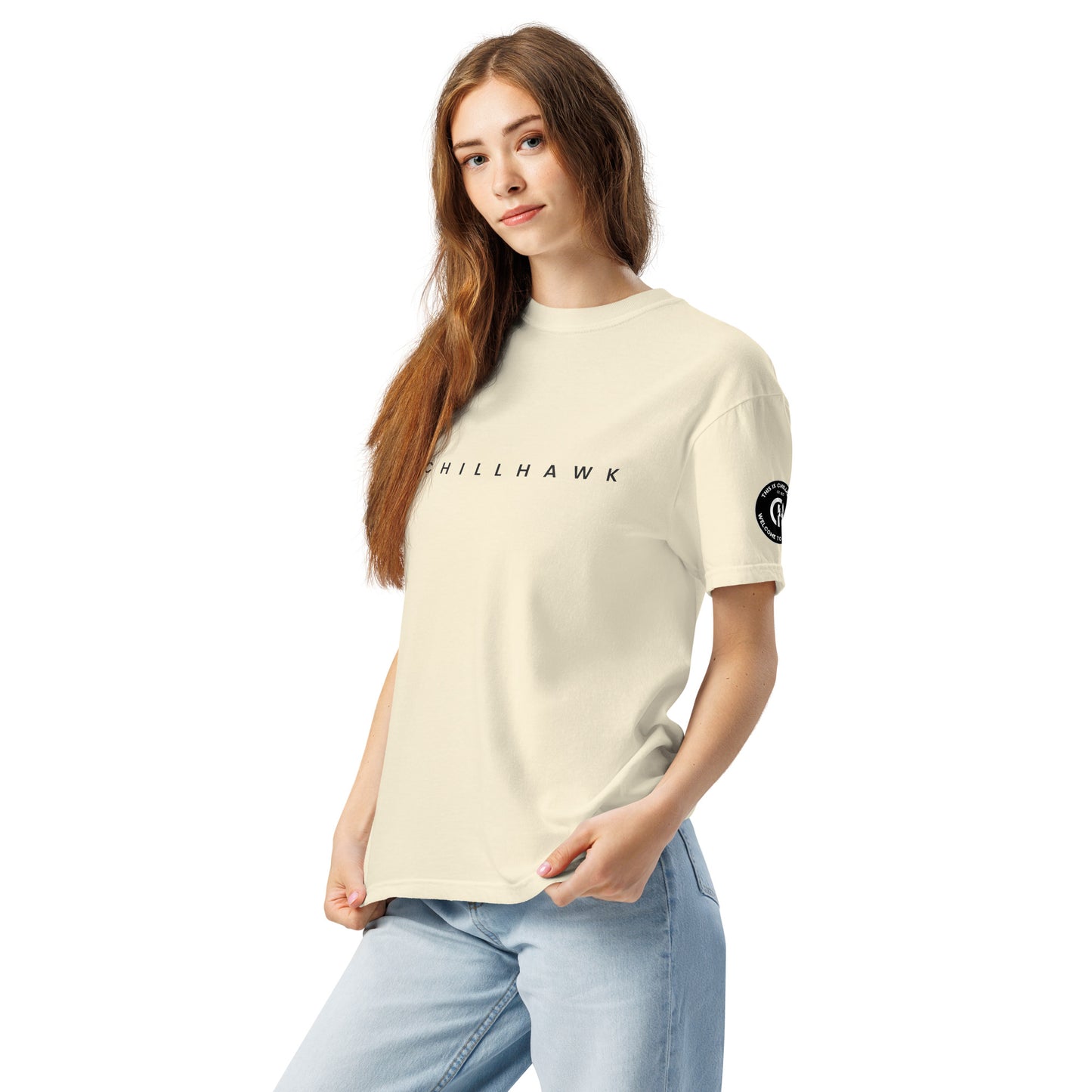 Chillhawk unisex heavyweight short sleeve t-shirt in ivory - a lady with light skin and long brown hair, is
wearing the t-shirt and light blue jeans. The word "CHILLHAWK" is printed in black on the front of the shirt, and a circular logo is visible on the left sleeve.
