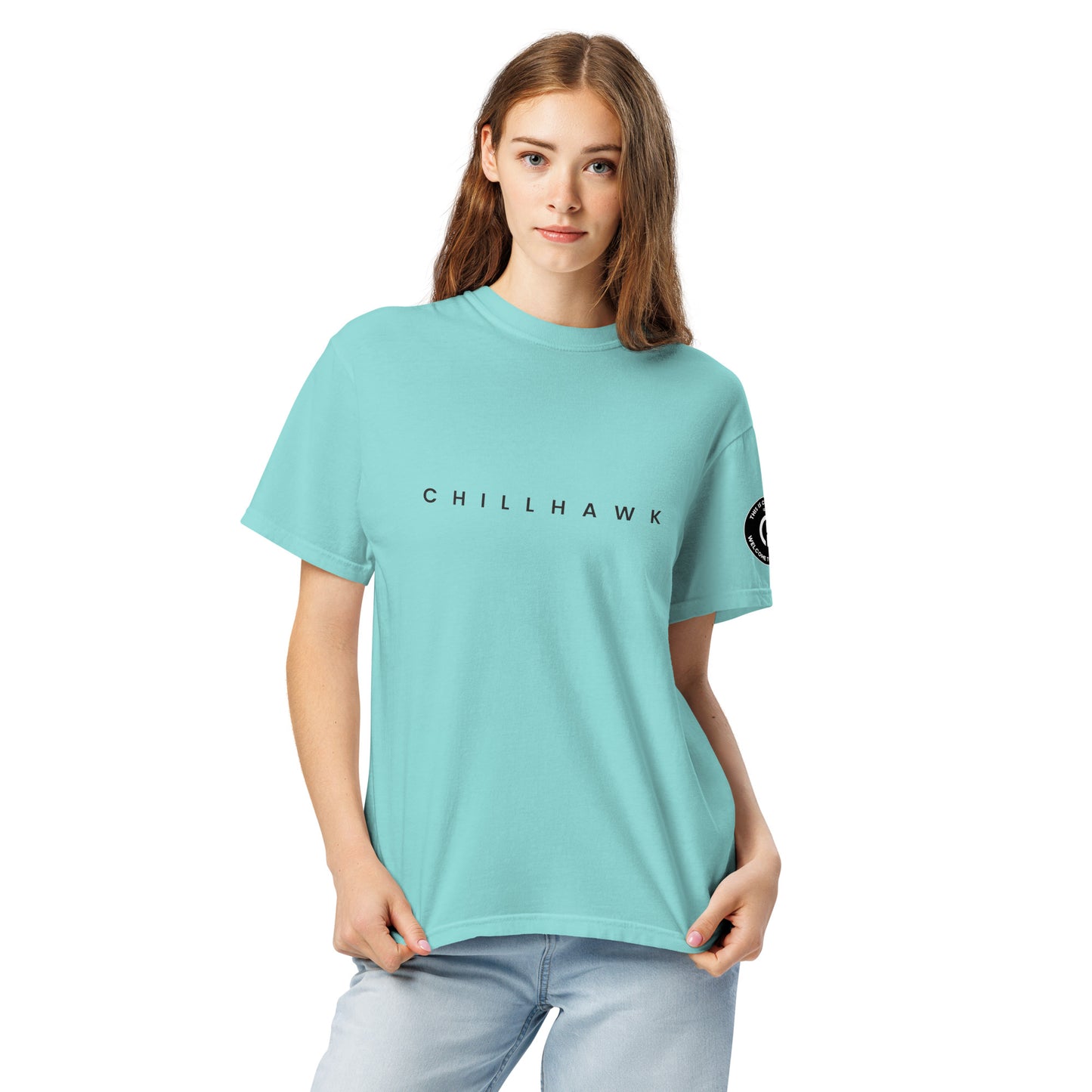Chillhawk unisex heavyweight short sleeve t-shirt in lagoon blue - a lady with light skin and long brown hair, is
wearing the t-shirt and light blue jeans. The word "CHILLHAWK" is printed in black on the front of the shirt, and a circular logo is visible on the left sleeve.