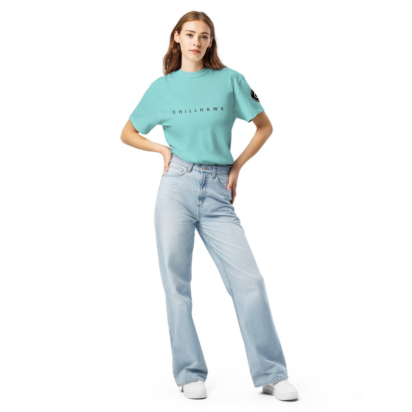 Chillhawk unisex heavyweight short sleeve t-shirt in lagoon blue - a lady with light skin and long brown hair, is standing with hands on hips wearing the t-shirt, light blue jeans, and white sneakers. The word "CHILLHAWK" is printed in black on the front of the shirt, and a circular logo is visible on the left sleeve.