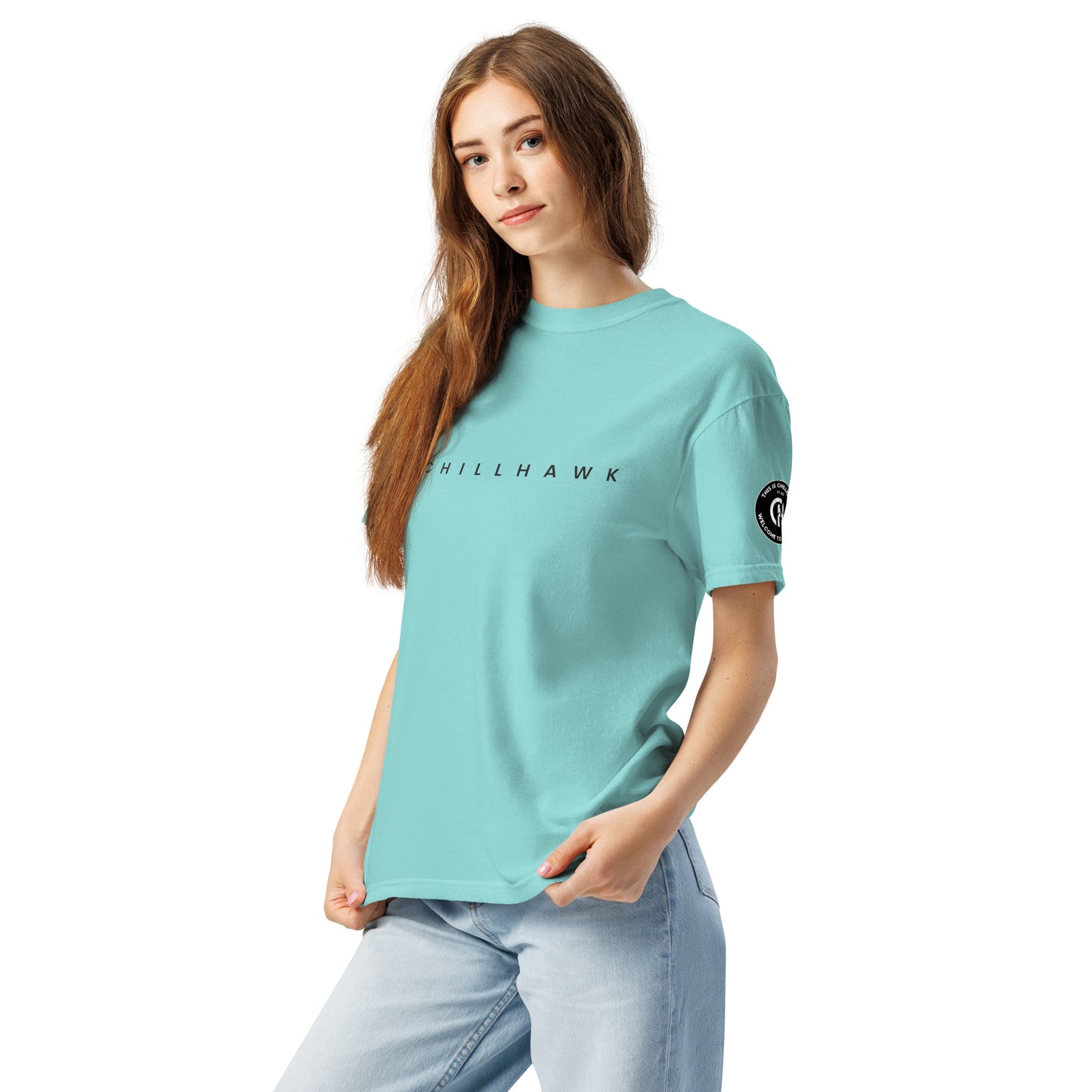 Chillhawk unisex heavyweight short sleeve t-shirt in lagoon blue - a lady with light skin and long brown hair, is
wearing the t-shirt and light blue jeans. The word "CHILLHAWK" is printed in black on the front of the shirt, and a circular logo is visible on the left sleeve.