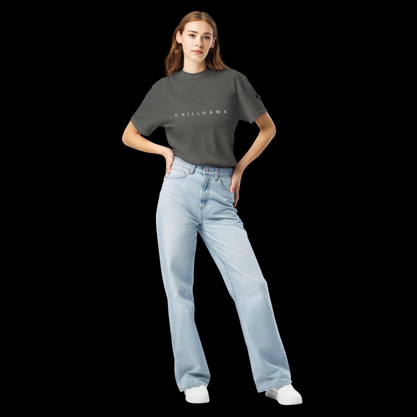 Chillhawk unisex heavyweight short sleeve t-shirt in pepper - front view of a lady with light skin and long brown hair, is wearing the t-shirt and light blue jeans. The word "CHILLHAWK" is printed in black on the front of the shirt, and a circular logo is visible on the left sleeve.
