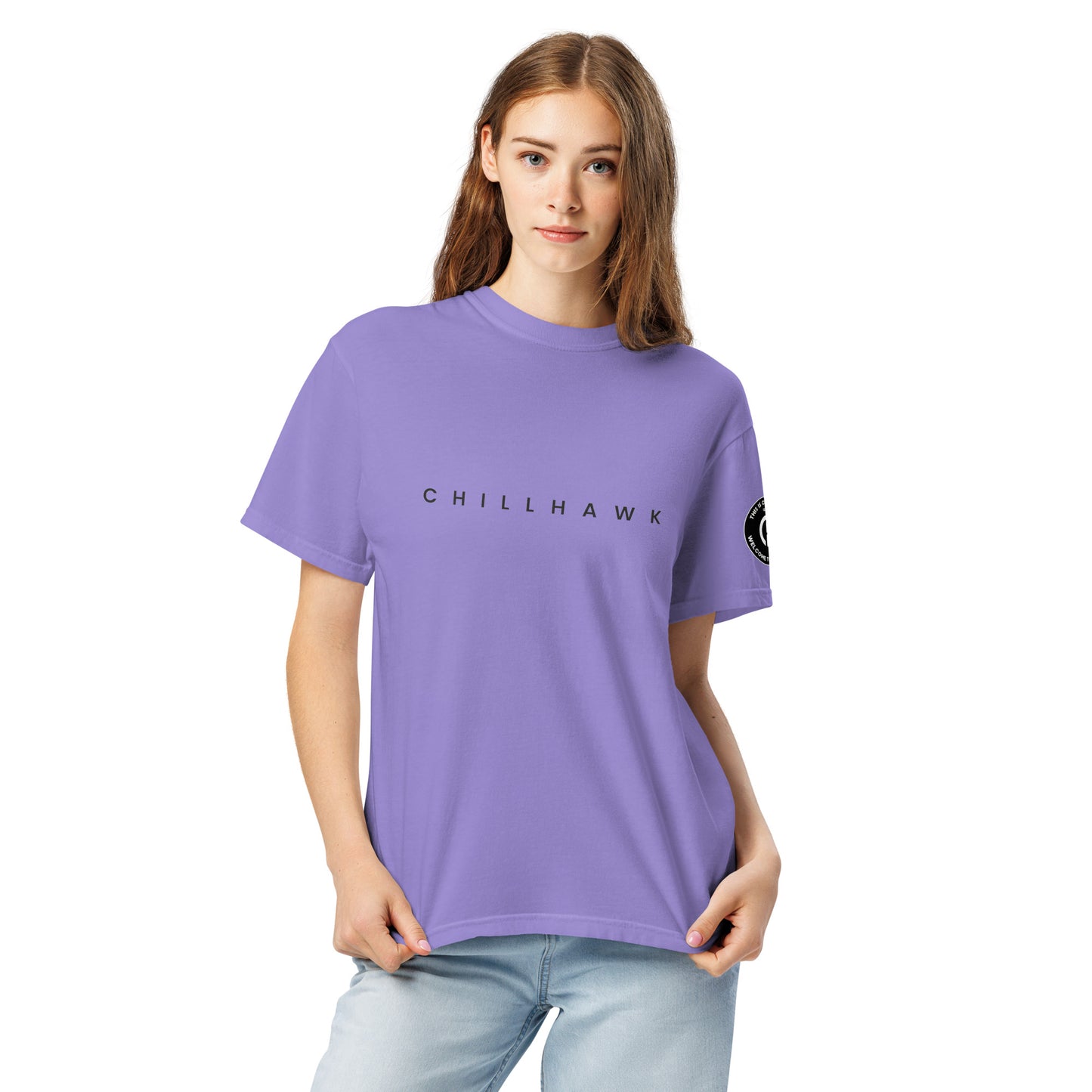 Chillhawk unisex heavyweight short sleeve t-shirt in violet - a lady with light skin and long brown hair, is
wearing the t-shirt and light blue jeans. The word "CHILLHAWK" is printed in black on the front of the shirt, and a circular logo is visible on the left sleeve.