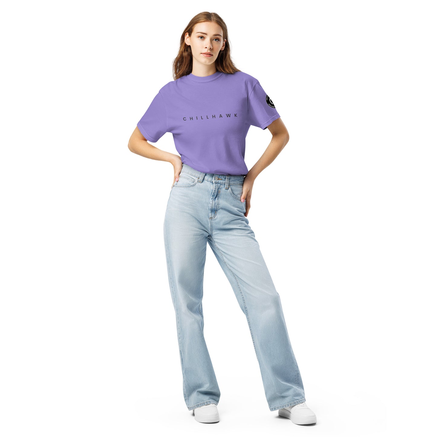 Chillhawk unisex heavyweight short sleeve t-shirt in violet - a lady with light skin and long brown hair, is standing with hands on hips wearing the t-shirt, light blue jeans, and white sneakers. The word "CHILLHAWK" is printed in black on the front of the shirt, and a circular logo is visible on the left sleeve.