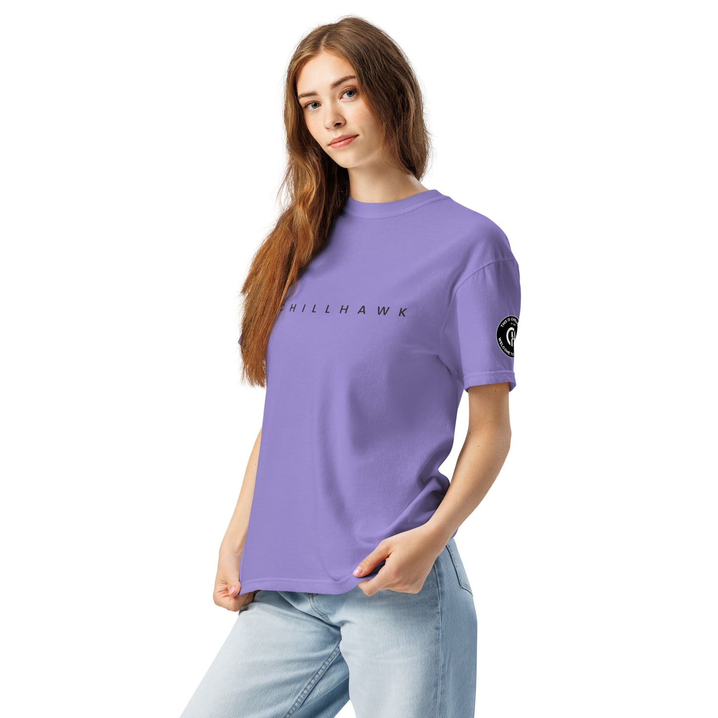 Chillhawk unisex heavyweight short sleeve t-shirt in violet - a lady with light skin and long brown hair, is
wearing the t-shirt and light blue jeans. The word "CHILLHAWK" is printed in black on the front of the shirt, and a circular logo is visible on the left sleeve.