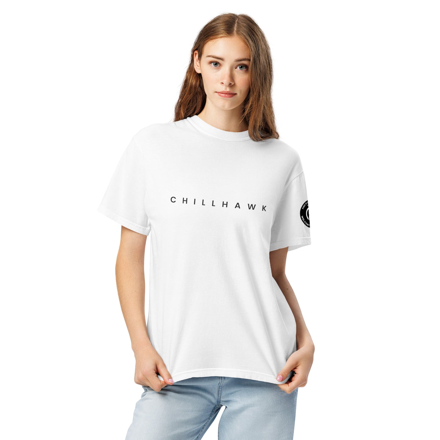 Chillhawk unisex heavyweight short sleeve t-shirt in white - front view of a lady with light skin and long brown hair, is wearing the t-shirt and light blue jeans. The word "CHILLHAWK" is printed in black on the front of the shirt, and a circular logo is visible on the left sleeve.
