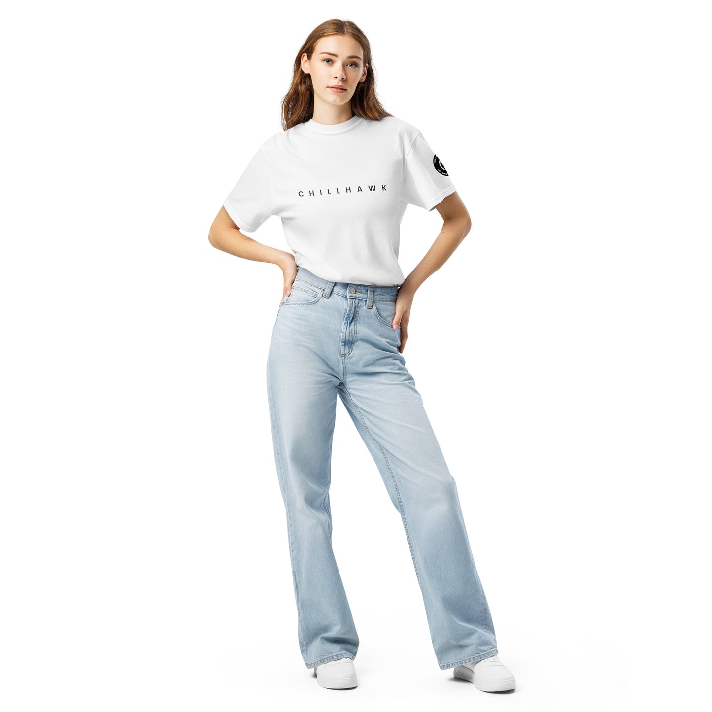 Chillhawk unisex heavyweight short sleeve t-shirt in white - front view of a lady with light skin and long brown hair, is wearing the t-shirt and light blue jeans. The word "CHILLHAWK" is printed in black on the front of the shirt, and a circular logo is visible on the left sleeve.