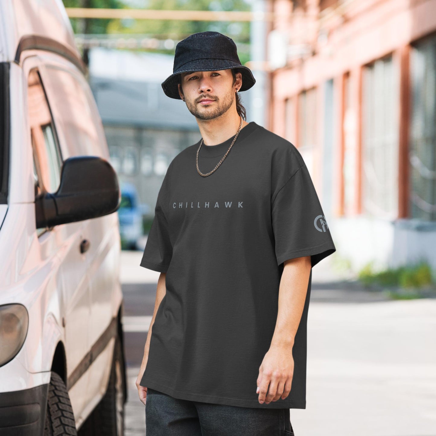 Chillhawk oversized faded t-shirt in faded black - a person with light skin, a short beard, and dark hair, is wearing the t-shirt, a black bucket hat, and a silver chain necklace. The word "CHILLHAWK" is printed in a lighter color on the chest, and a circular logo is on the left sleeve. The person is standing outdoors next to a white van.