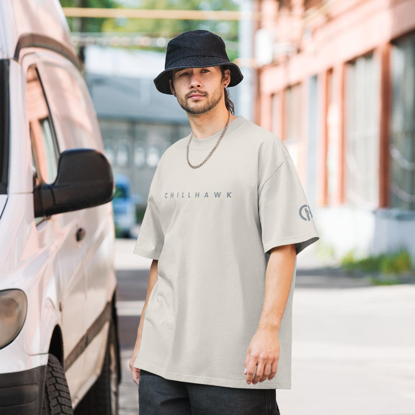 Chillhawk oversized faded t-shirt in faded bone - a person with light skin, a short beard, and dark hair, is wearing the t-shirt, a black bucket hat, and a silver chain necklace. The word "CHILLHAWK" is printed in a lighter color on the chest, and a circular logo is on the left sleeve. The person is standing outdoors next to a white van.
