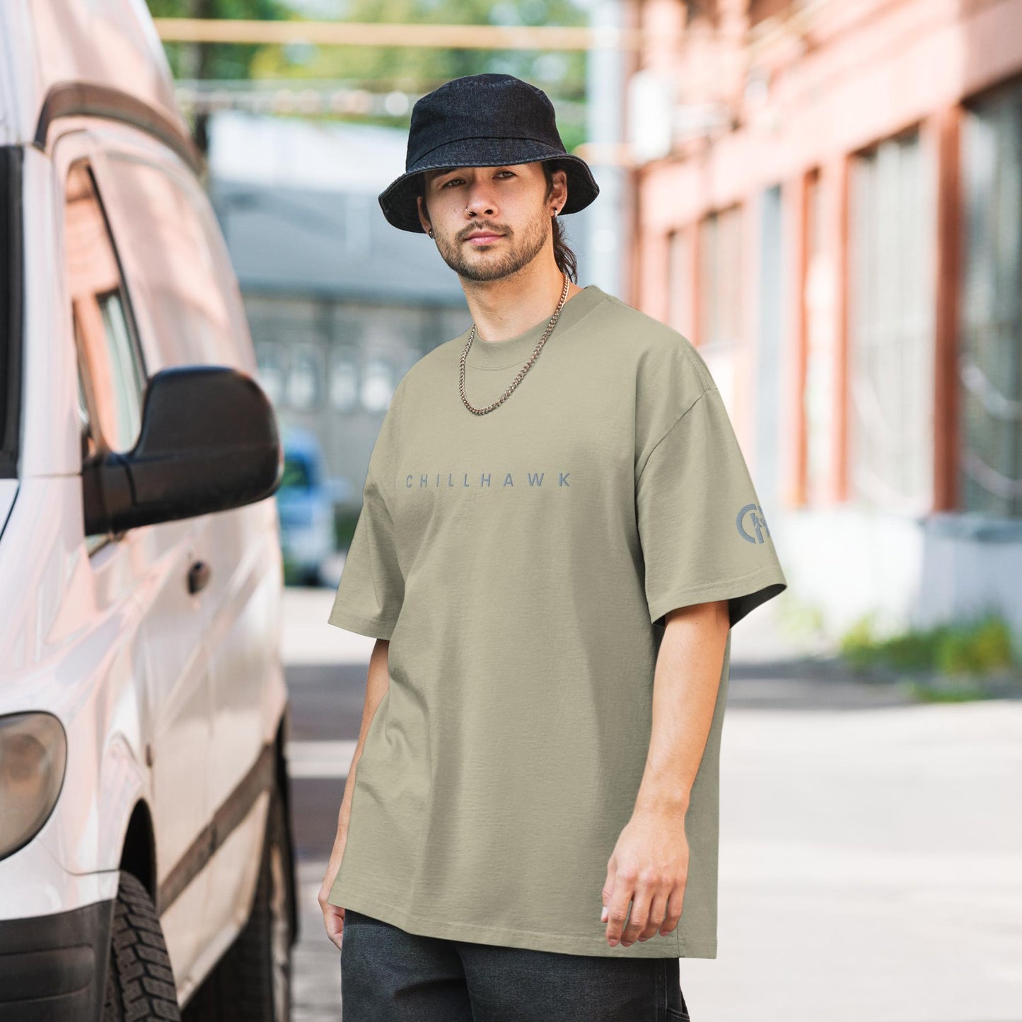Chillhawk oversized faded t-shirt in faded eucalyptus - a person with light skin, a short beard, and dark hair, is wearing the t-shirt, a black bucket hat, and a silver chain necklace. The word "CHILLHAWK" is printed in a lighter color on the chest, and a circular logo is on the left sleeve. The person is standing outdoors next to a white van.