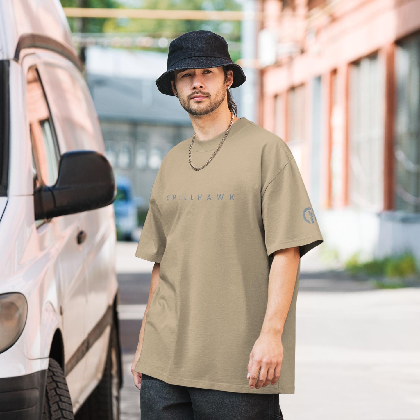 Chillhawk oversized faded t-shirt in faded khaki - a person with light skin, a short beard, and dark hair, is wearing the t-shirt, a black bucket hat, and a silver chain necklace. The word "CHILLHAWK" is printed in a lighter color on the chest, and a circular logo is on the left sleeve. The person is standing outdoors next to a white van.