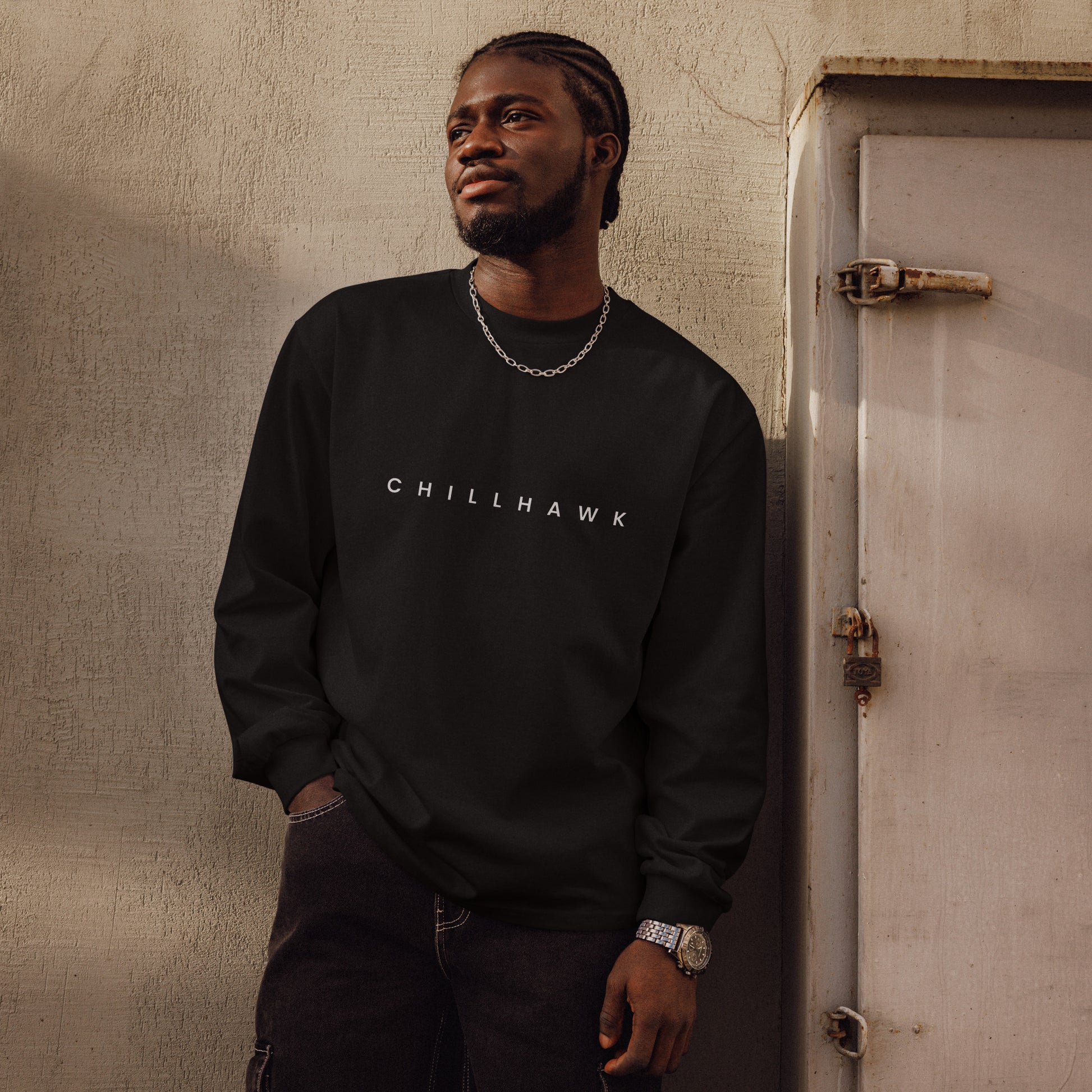 Chillhawk unisex premium heavyweight long sleeve shirt in black - a person with dark skin and braided hair, is shown looking to the side wearing the shirt and black jeans. The word "CHILLHAWK" is printed in white on the front of the shirt.