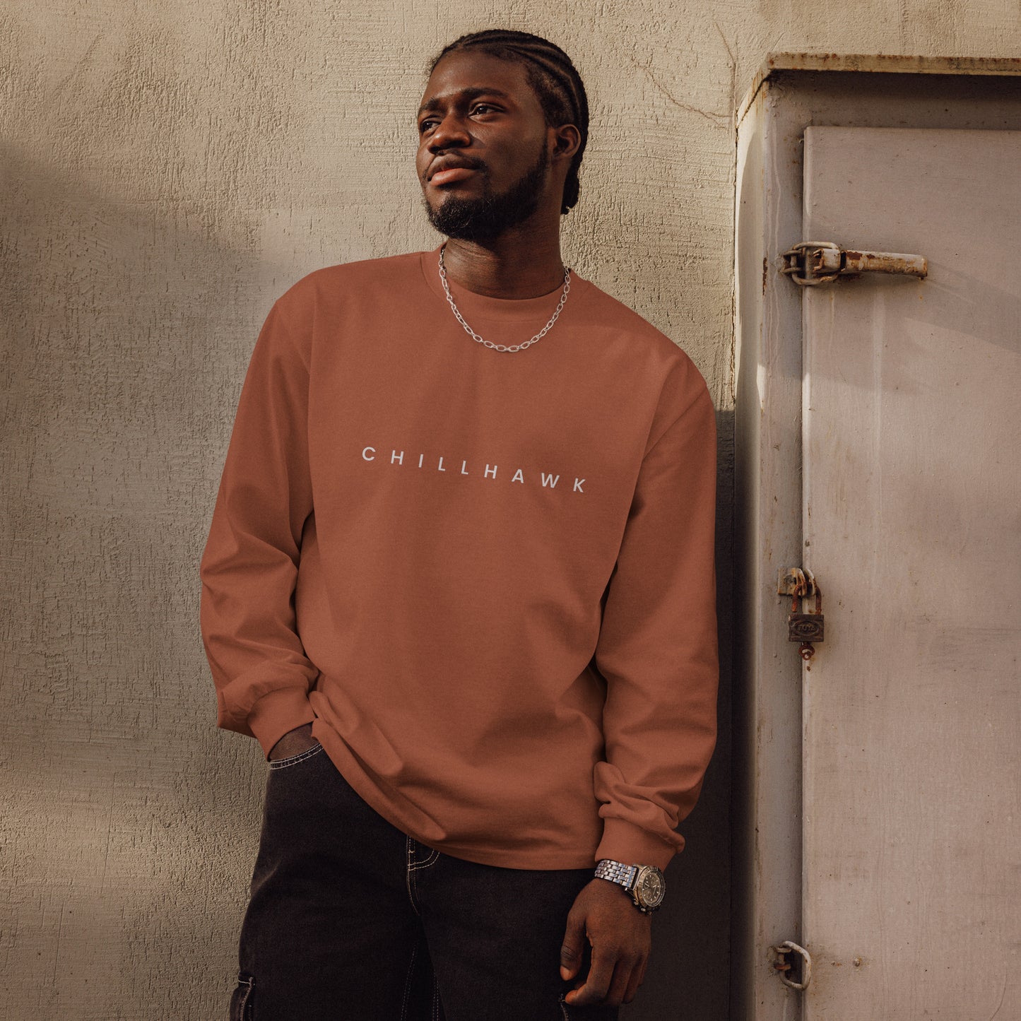 Chillhawk unisex premium heavyweight long sleeve shirt in clay - a person with dark skin and braided hair, is shown looking to the side wearing the shirt and black jeans. The word "CHILLHAWK" is printed in white on the front of the shirt.