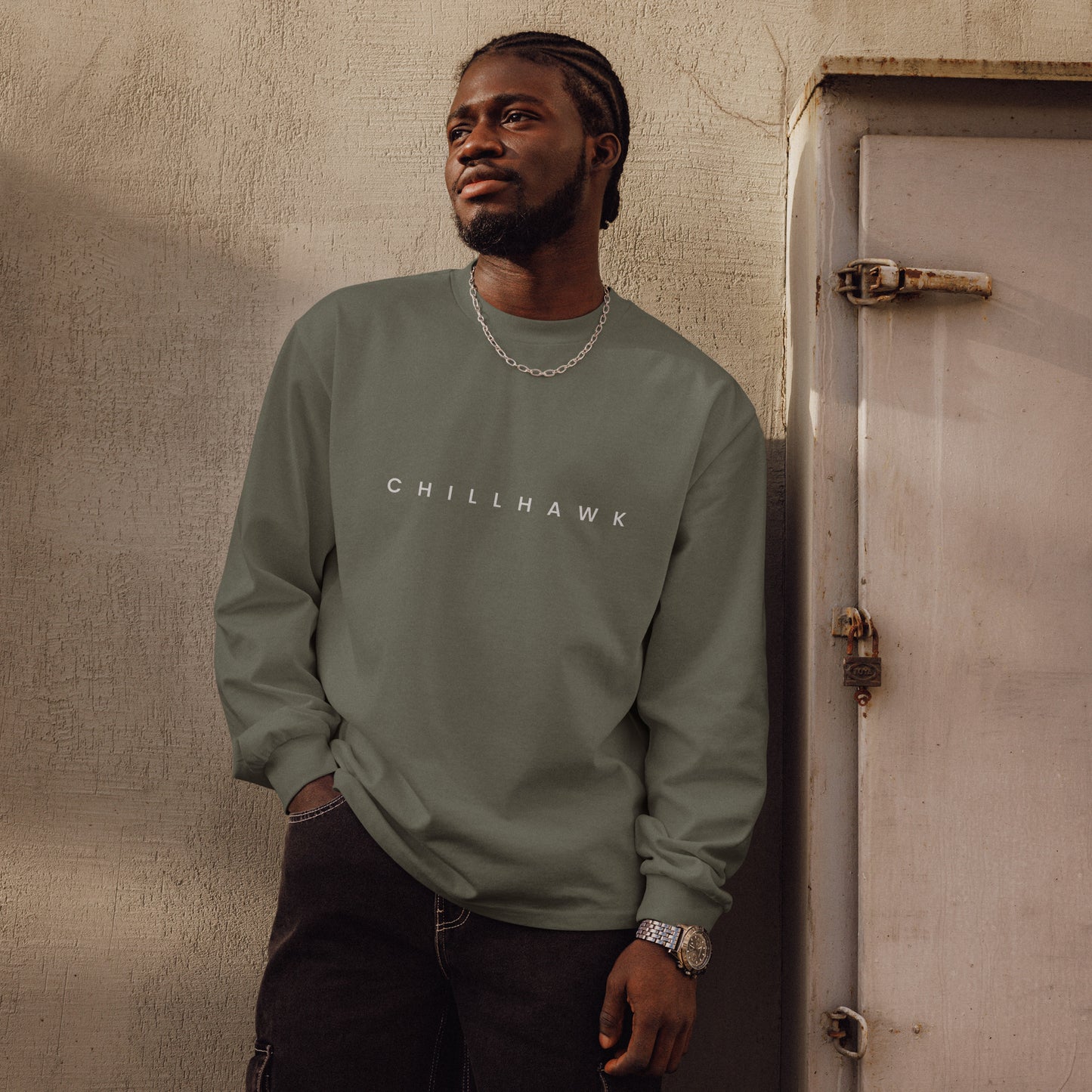 Chillhawk unisex premium heavyweight long sleeve shirt in cypress - a person with dark skin and braided hair, is shown looking to the side wearing the shirt and black jeans. The word "CHILLHAWK" is printed in white on the front of the shirt.